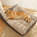 Dog lying on a large quilted pet bed in a room with wooden flooring and white curtains.